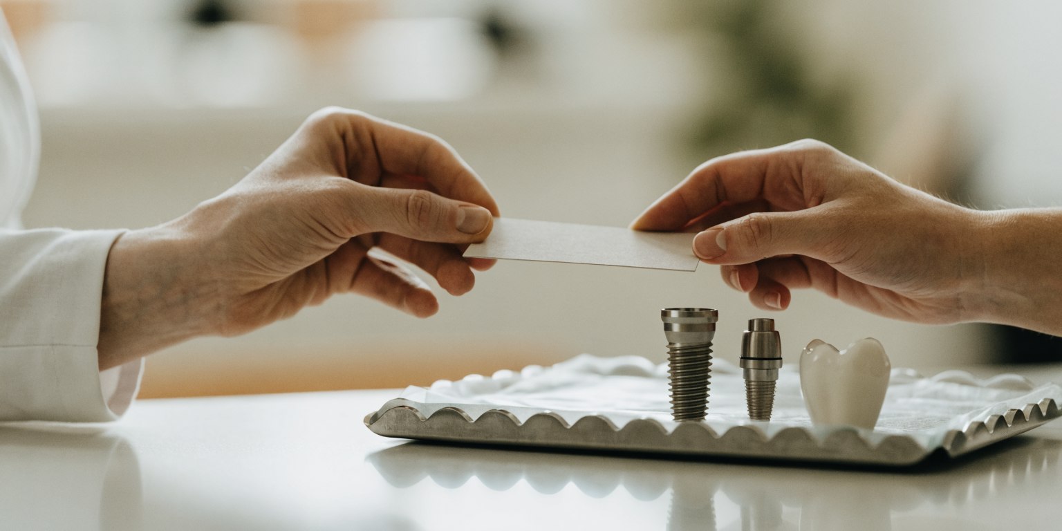Dentist handing a patient a written implant record beside implant, abutment and crown components